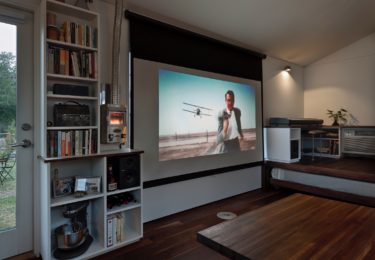 The large window array next to the door is conveniently sized in 9:16 proportion, and a projector screen hanging above it doubles as a shade. A small LED projector affixed near the bookcase displays a 90” image, accompanied by speakers on either side of the window. A Dickenson marine propane heater appears to the left of the screen.